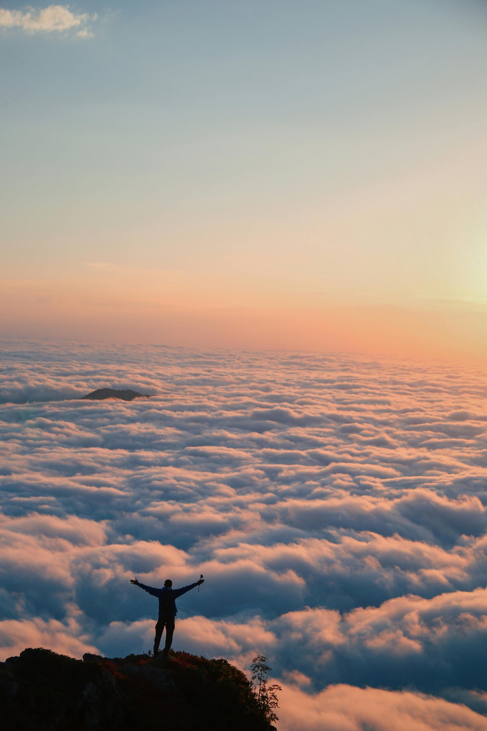 A person stands triumphantly on a cliff edge, surrounded by endless clouds at sunrise.